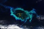 A phytoplankton bloom in the southern Pacific Ocean appears as an oval-shaped area of green and chalky blue water surrounding a group of small islands.