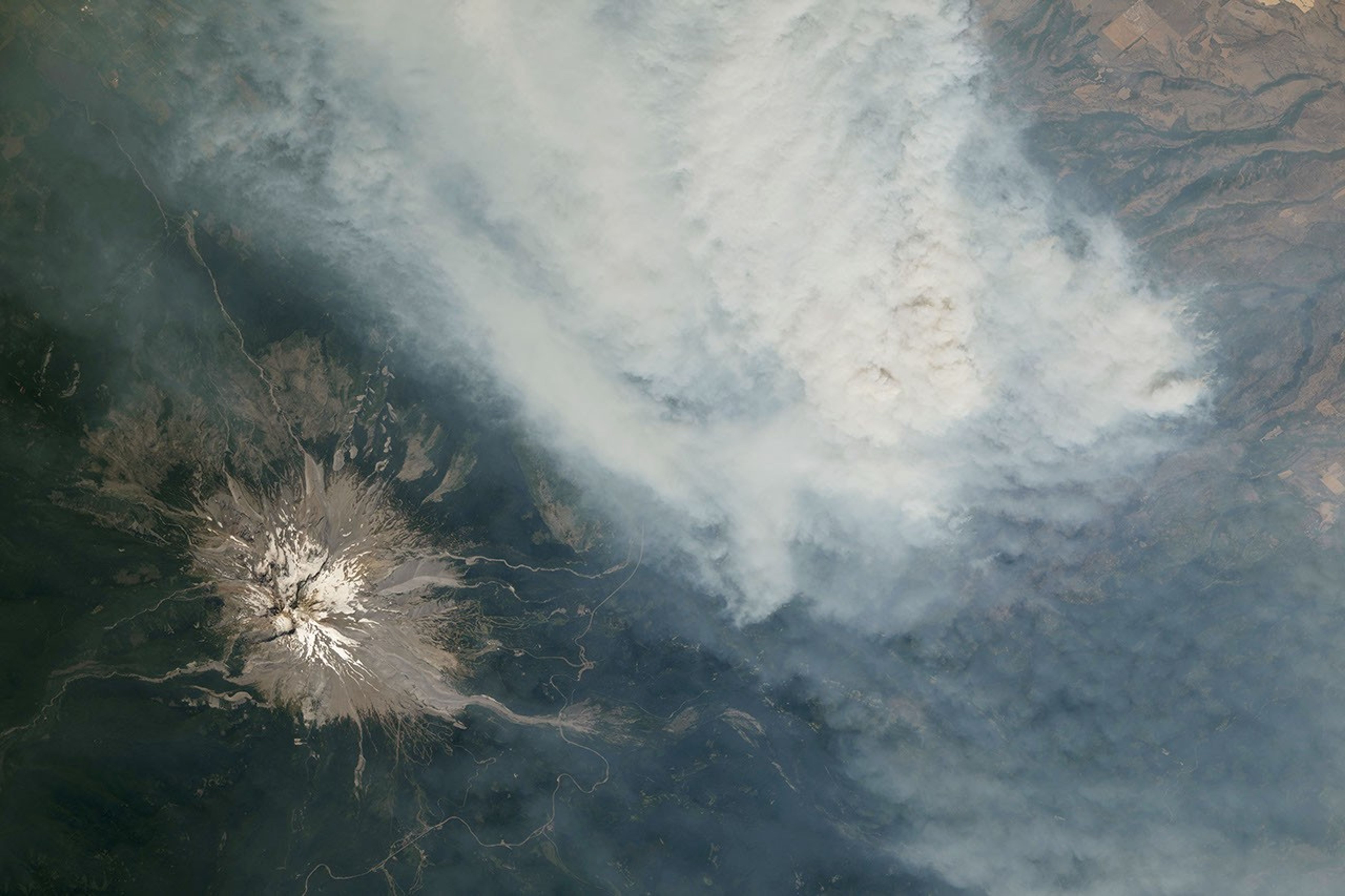 An ice-capped volcano stands in the bottom-left corner, with a wide plume of brown-gray smoke filling most of the rest of the frame.