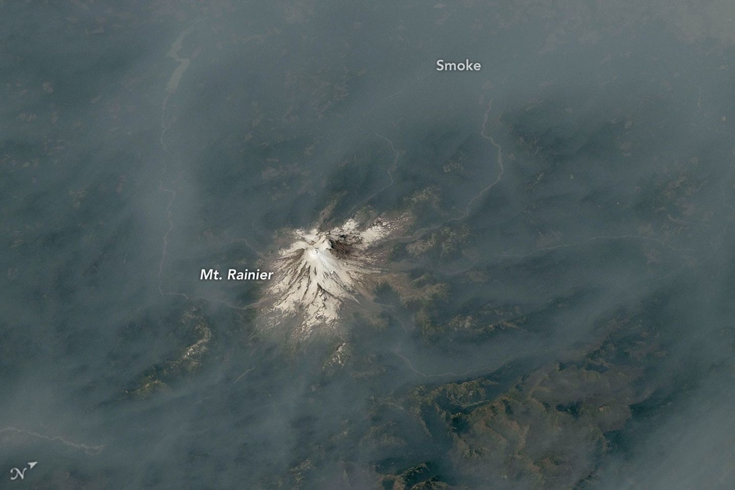 An ice-capped volcano stands at the center of the image, its surrounding terrain blanketed in brown-gray smoke.