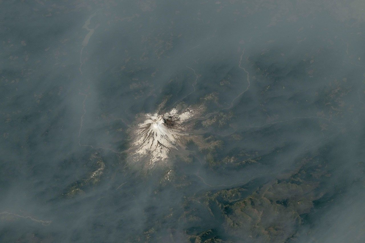 An ice-capped volcano stands at the center of the image, its surrounding terrain blanketed in brown-gray smoke.