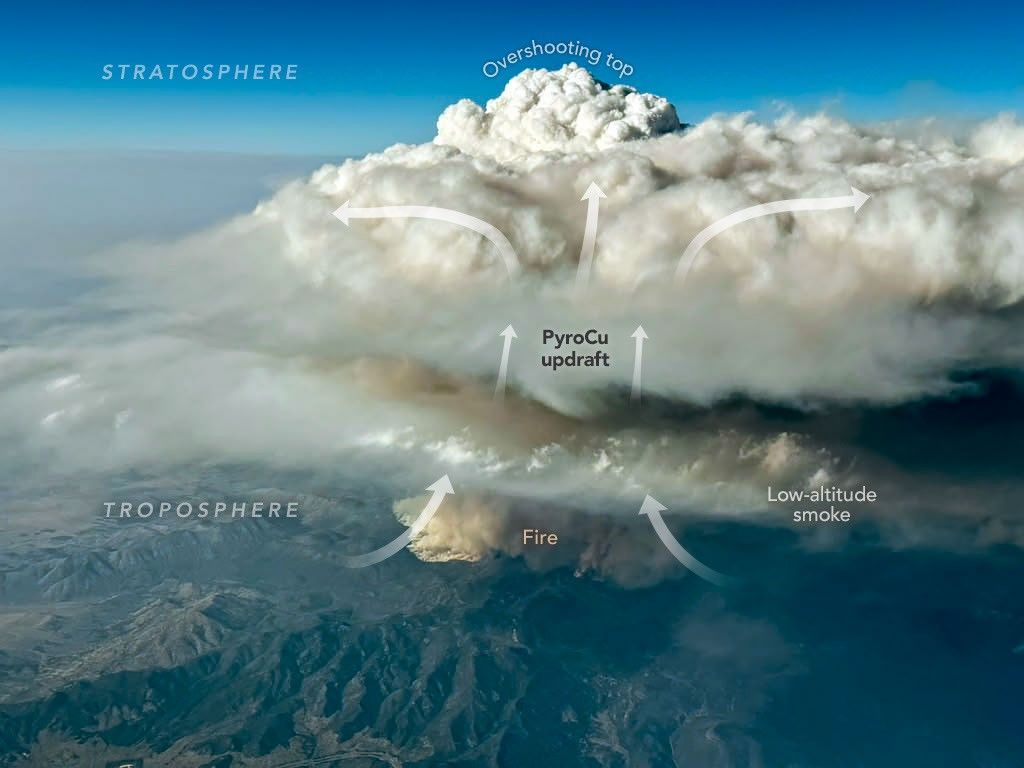 An aerial image shows a thick, puffy white cloud rising high above a patch of darker smoke visible near a surface of variable mountainous terrain.