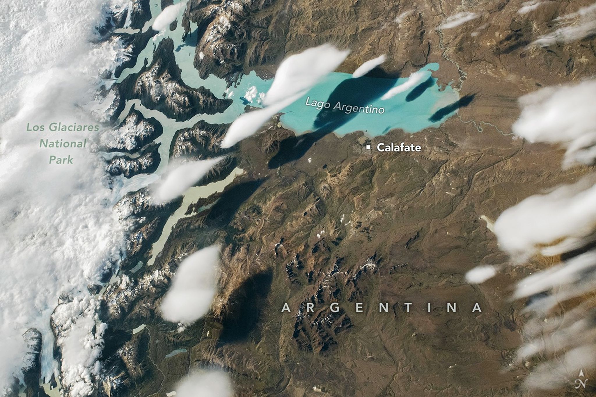 Several narrow, white clouds resembling fish are visible over the teal waters of Lago Argentino. The landscape surrounding the lake is brown and barren. The left side of the image includes a label for Los Glaciares National Park.