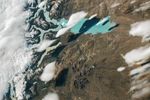 Several narrow, white clouds resembling fish are visible over the teal waters of Lago Argentino. The landscape surrounding the lake is brown and barren. The left side of the image includes a label for Los Glaciares National Park.