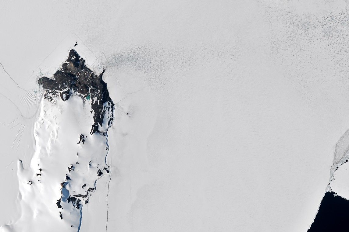 A satellite image shows sea ice along Antarctica’s Ross Island near McMurdo Station. Buildings and fuel tanks are visible on land. The start of a straight channel, cut by an icebreaker through the sea ice, is visible toward the right, originating from small area of dark open water.