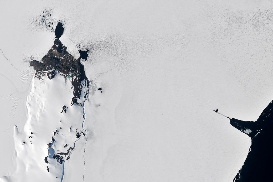A satellite image shows sea ice along Antarctica’s Ross Island near McMurdo Station. Buildings and fuel tanks are visible on land. The start of a straight channel, cut by an icebreaker through the sea ice, is visible toward the right, originating from small area of dark open water.