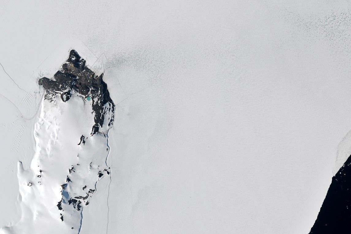 A satellite image shows sea ice along Antarctica’s Ross Island near McMurdo Station. Buildings and fuel tanks are visible on land. The start of a straight channel, cut by an icebreaker through the sea ice, is visible toward the right, originating from small area of dark open water.