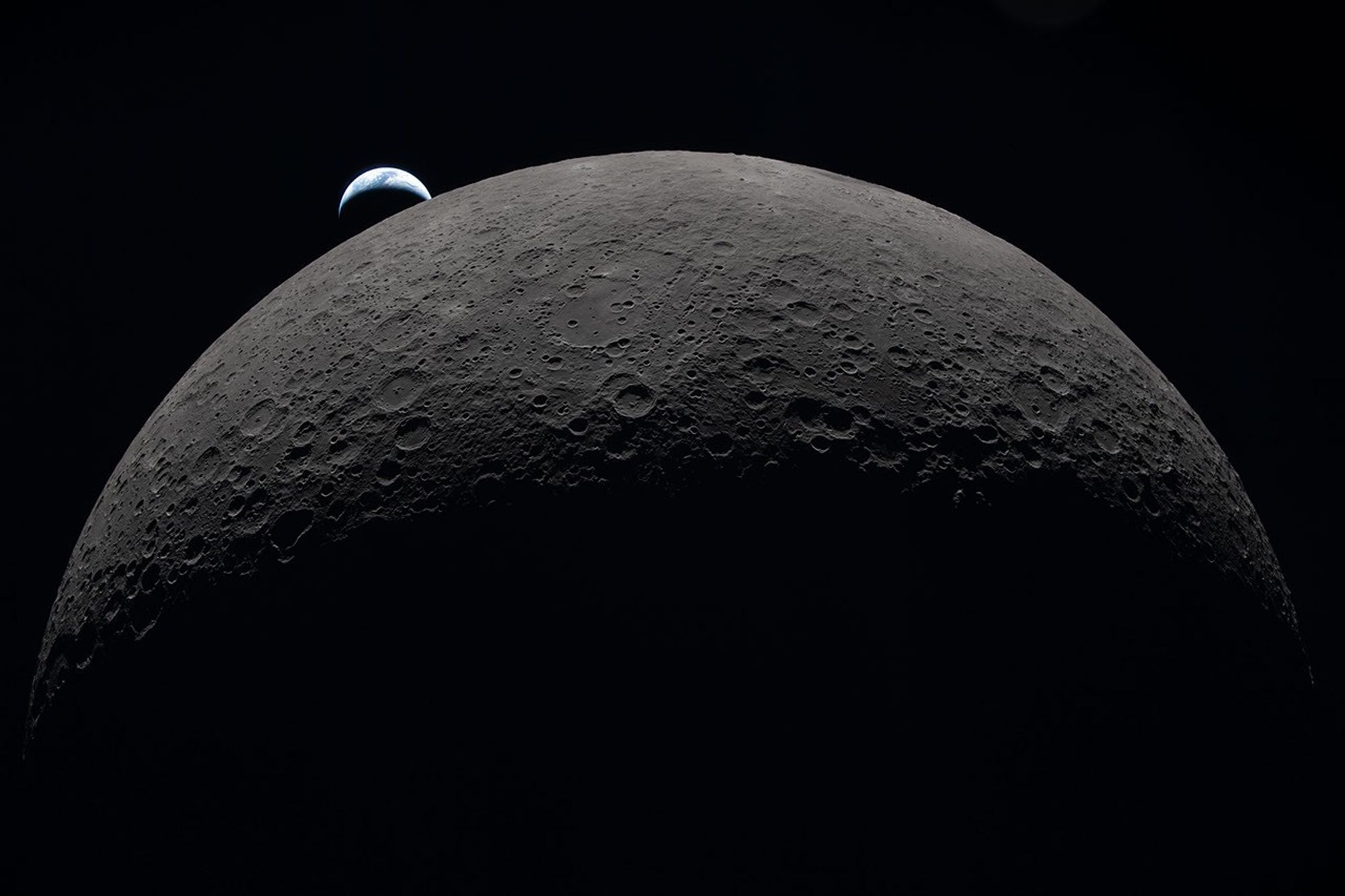 The gray-brown, heavily cratered Moon dominates the frame against black space, with a partially lit crescent Earth setting behind its upper-left edge.