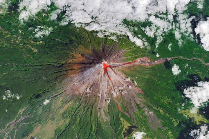 The upper slopes of Mayon volcano appear brown, with several narrow channels radiating from the crater. A red infrared heat signature appears near the summit, with red streaks extending east and southeast. The lower slopes are green and forested. Farmland and towns are visible in the lower part of the image.