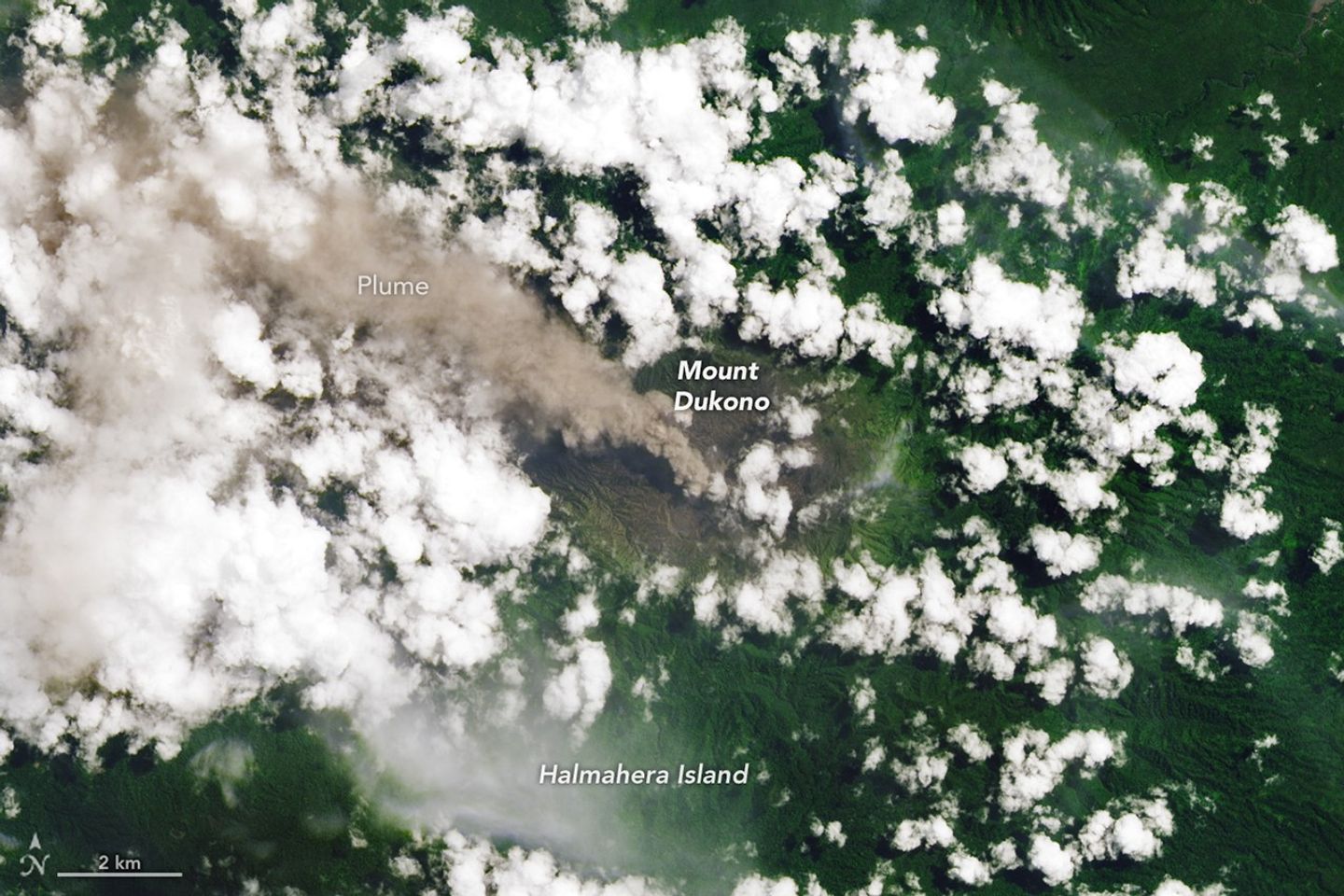 An ash-rich plume streams northwest from the volcano amidst scattered puffy, white clouds.