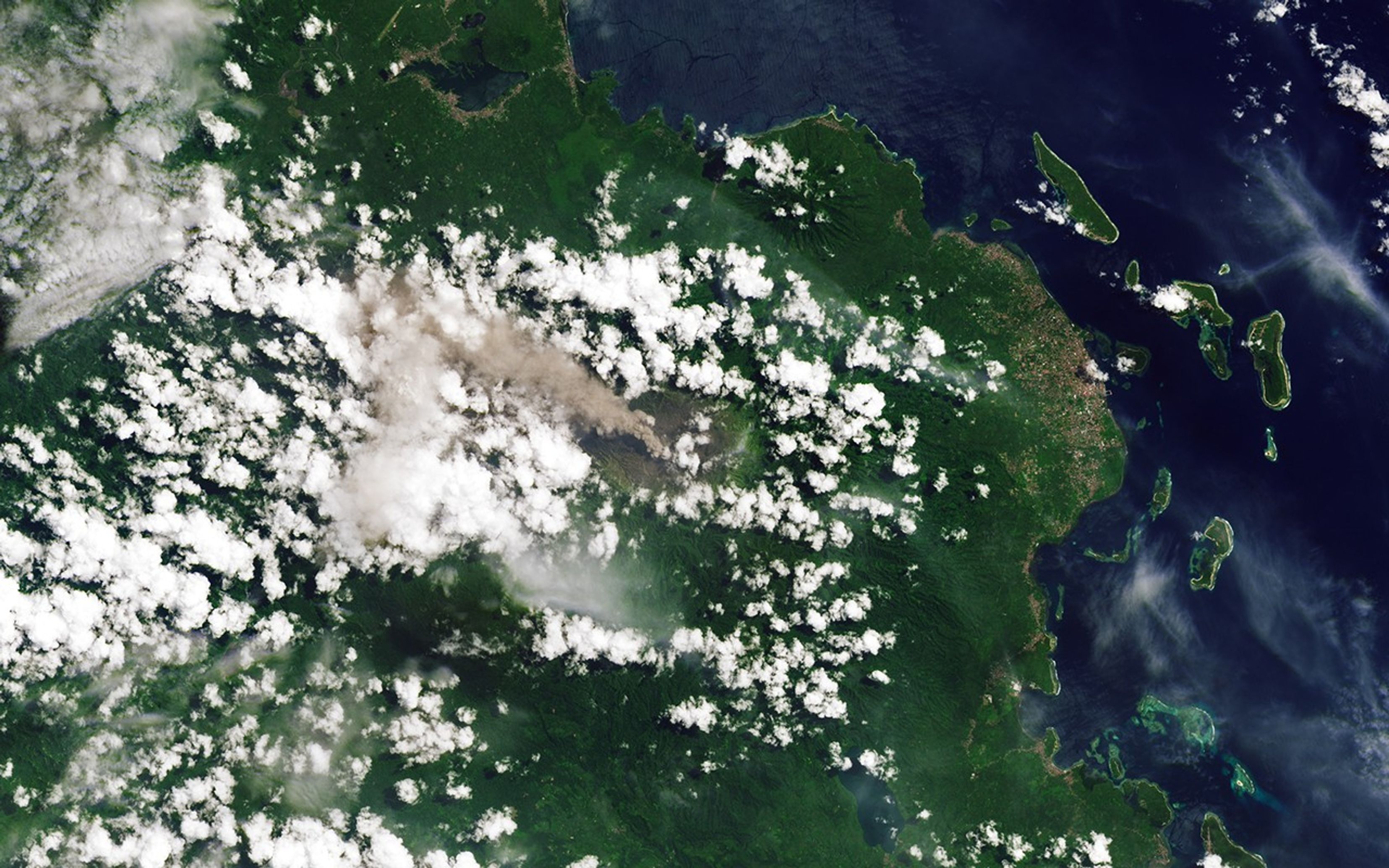 An ash-rich plume streams northwest from the volcano amidst scattered puffy, white clouds.