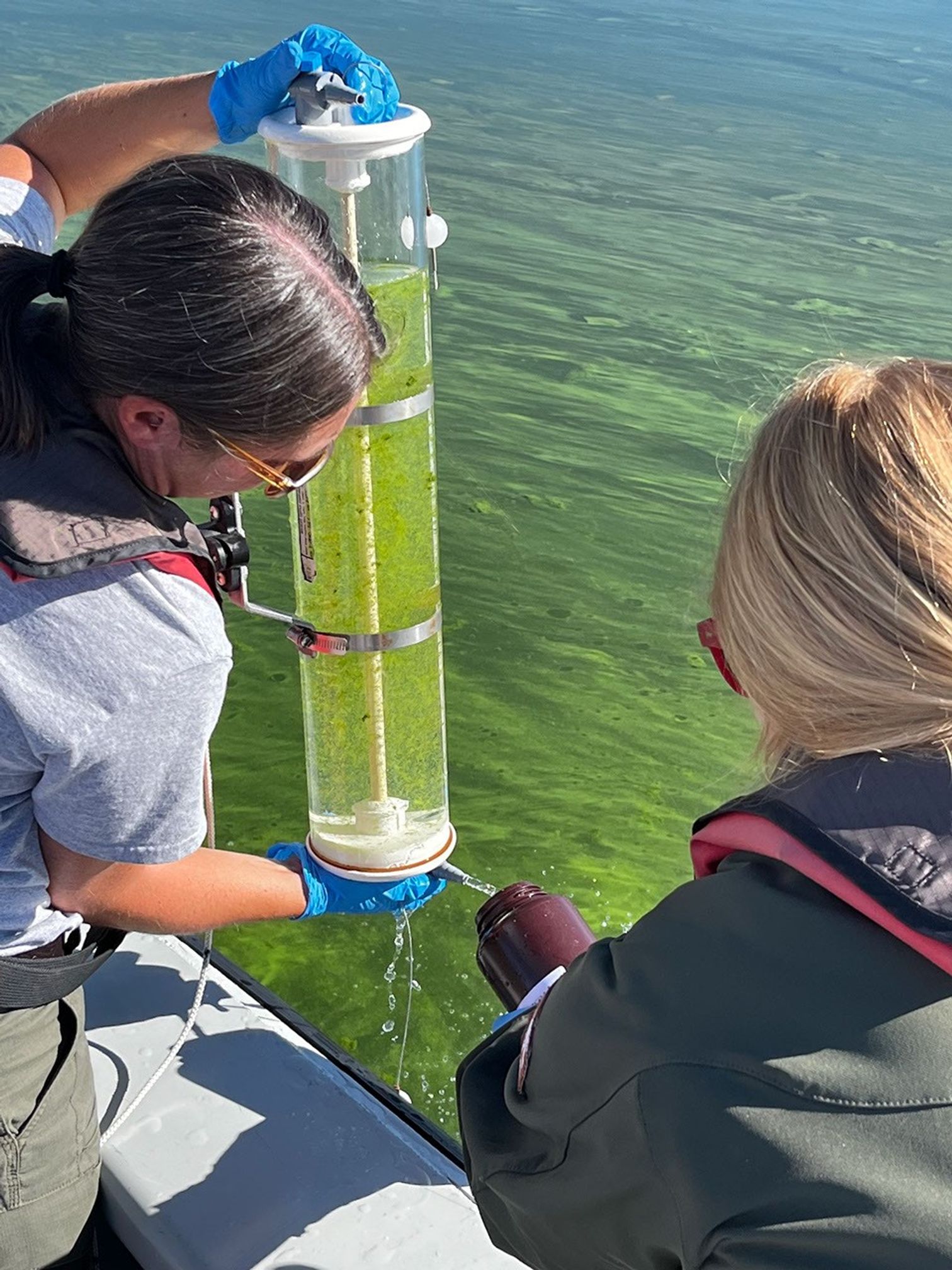A photograph shows two female researchers collecting green, algae-rich water in a cylindrical container.
