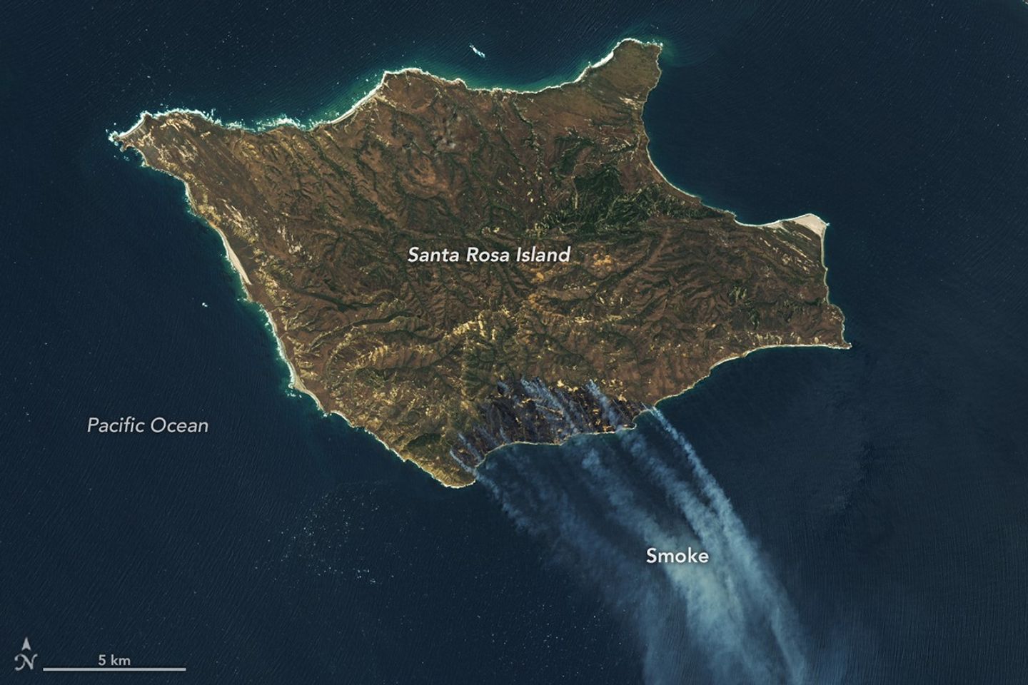 A downward-looking image of Santa Rosa Island is mostly brown, with a darker brown area on the bottom-right side. Gray-white smoke drifts toward the bottom-right over dark blue ocean water.