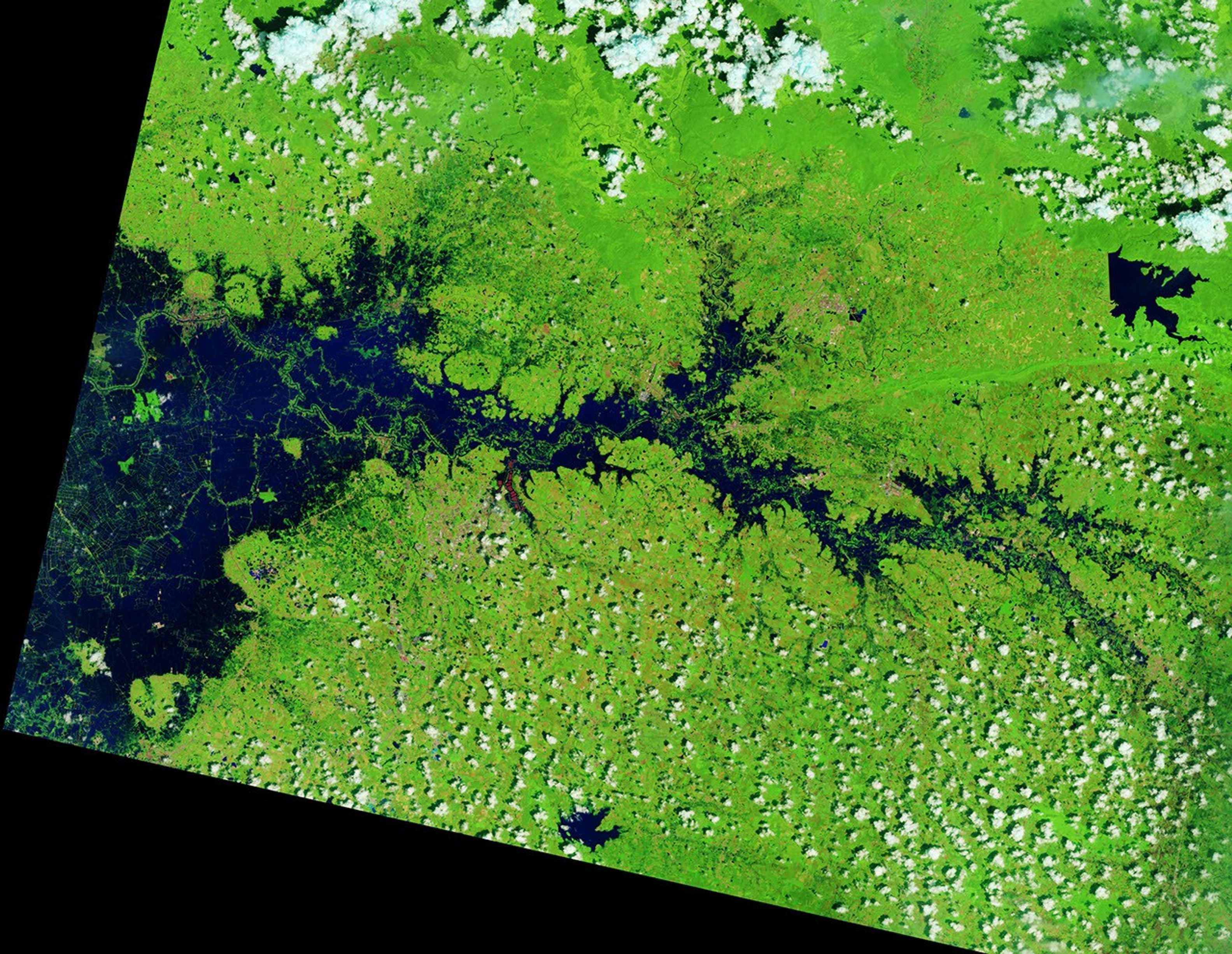 In a false-color satellite image of the same area, floodwaters (dark blue) flank a meandering river and cover a large swath of agricultural or aquacultural land primarily on the left side of the scene.
