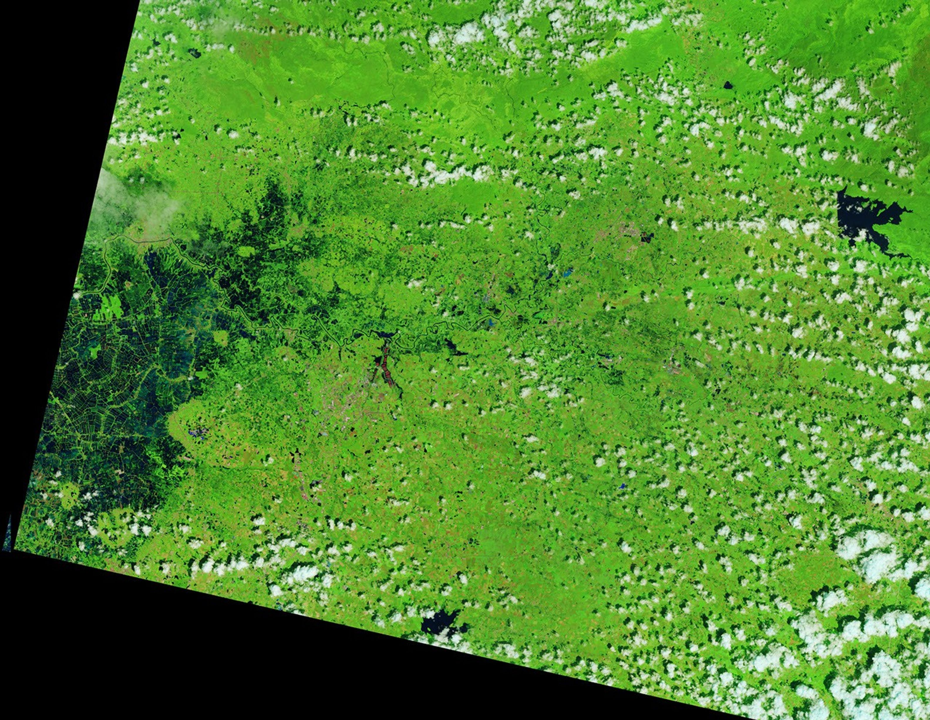 A narrow river (dark blue) meanders across vegetated land (bright green) in a false-color satellite image. Small geometric ponds or waterlogged fields appear on the left side of the scene.