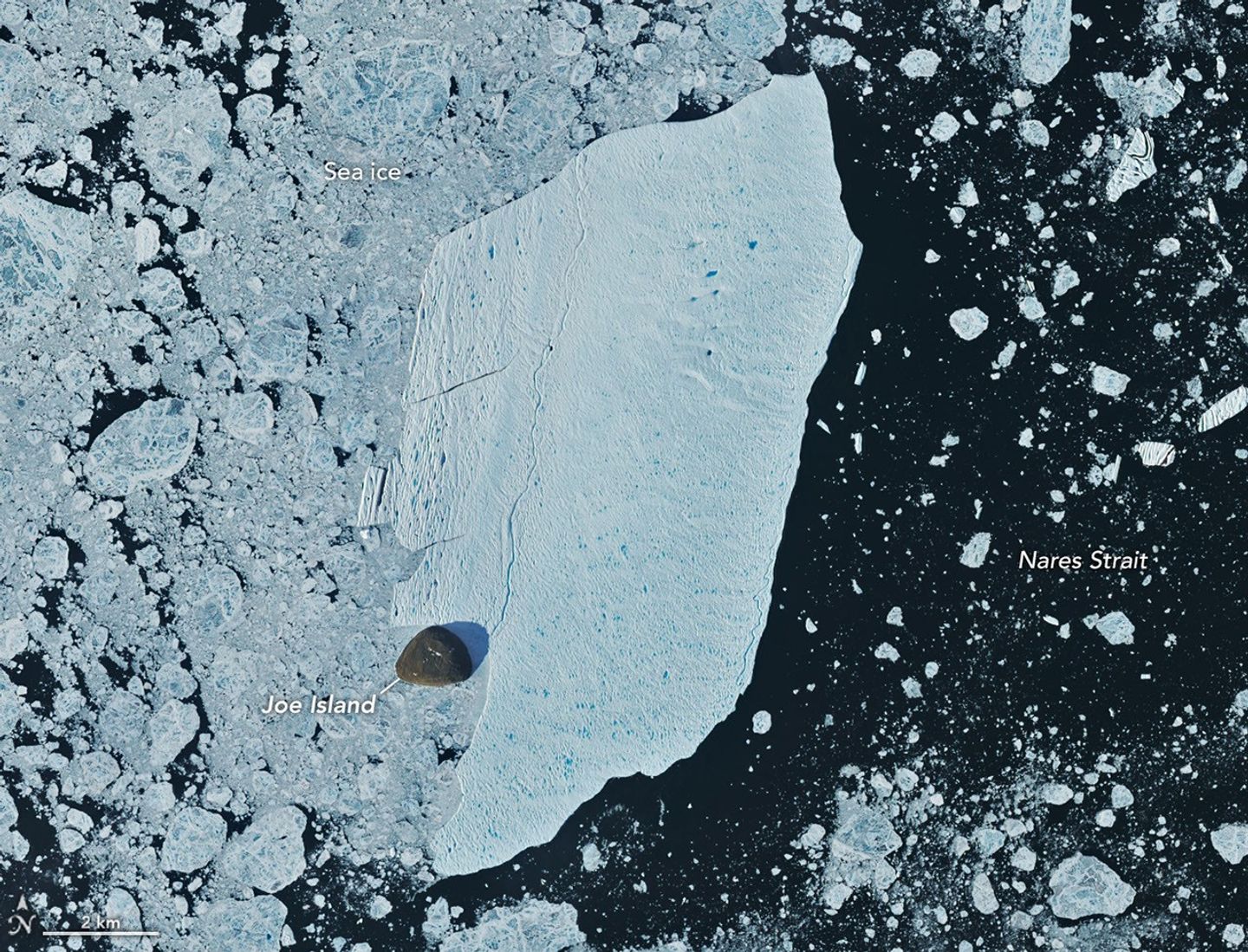 A detailed satellite view shows the iceberg wedged against the small, brown island, with sea ice packed densely to its left and more sparsely to its right.