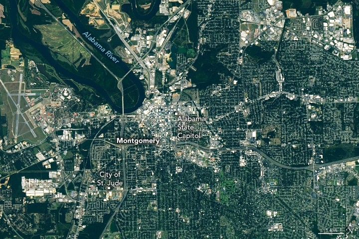 Montgomery is the focus of a satellite image that shows the city along a bend in the Alabama River. Labels mark the location of City of St. Jude on the southwestern outskirts of the city and the Alabama state capitol in the heart of the city. The most densely parts of the city appear gray while the city's outskirts are greener and have more tree cover.