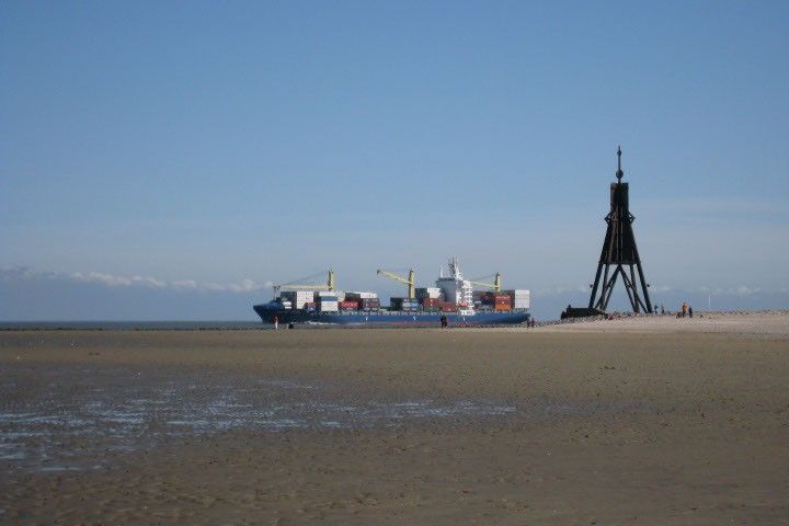 A broad tidal mudflat fills the foreground of this photo.  A sliver of deeper water is visible on the horizon, where a cargo ship loaded with shipping containers and cranes appears to be traveling to the left.