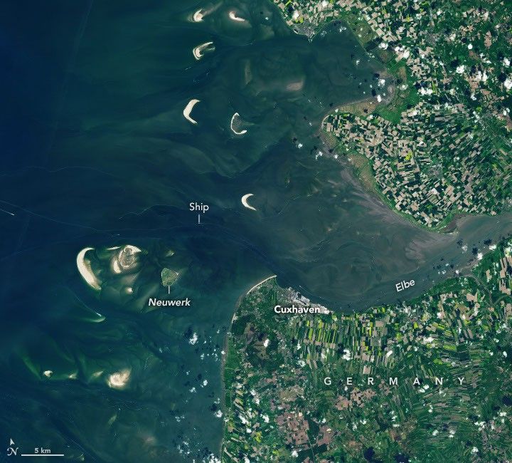 The Elbe river in Germany into the North Sea on the left side of the image. At high tide, a handful of small islands and crescent-shaped sandbars remain exposed above the water level near shore. Ships are visible as small, bright specks on the water.