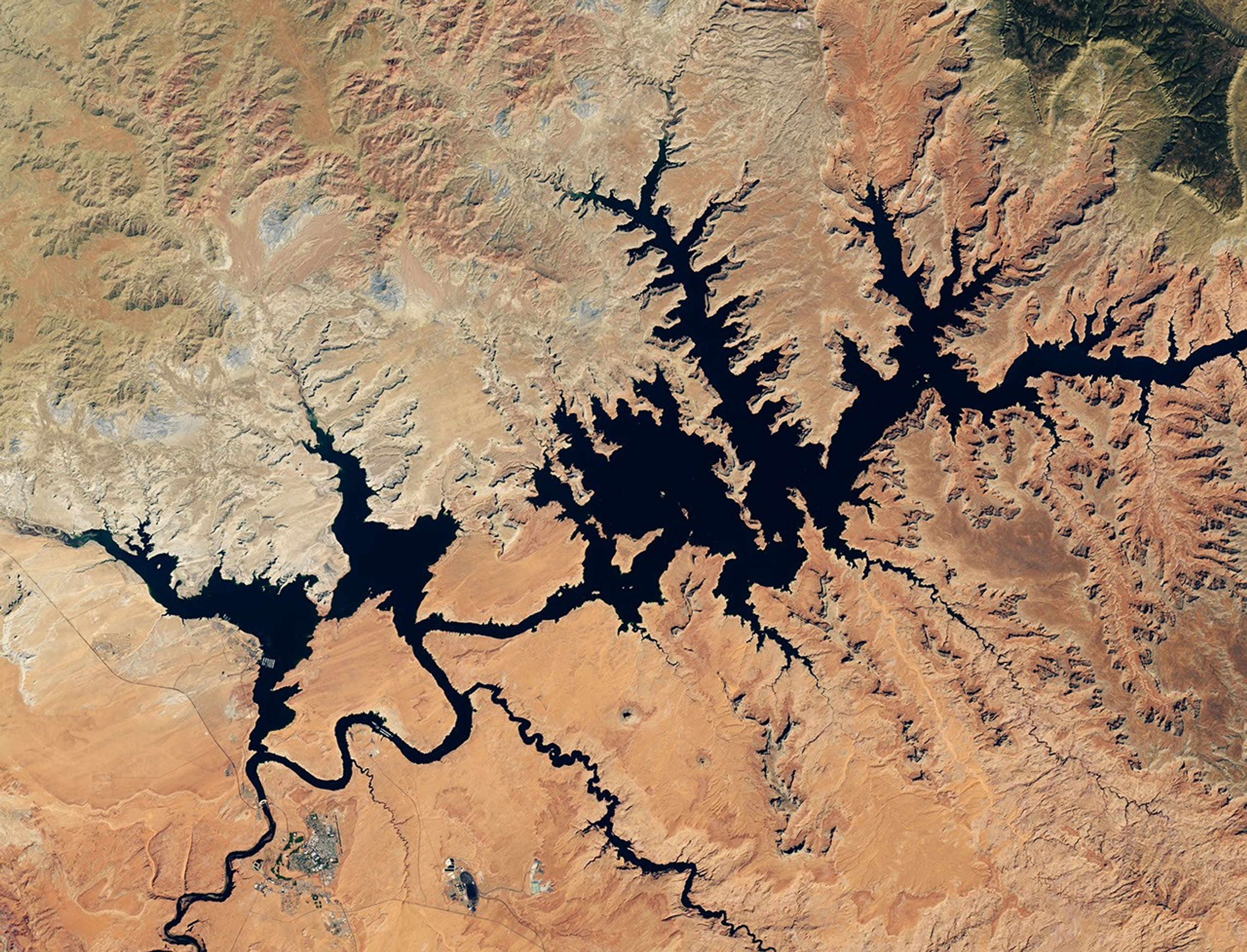 Dark blue water in Lake Powell fills several bays and branching canyons amid a light orange desert landscape. The Glen Canyon Dam and the town of Page, Arizona, are visible in the lower left.