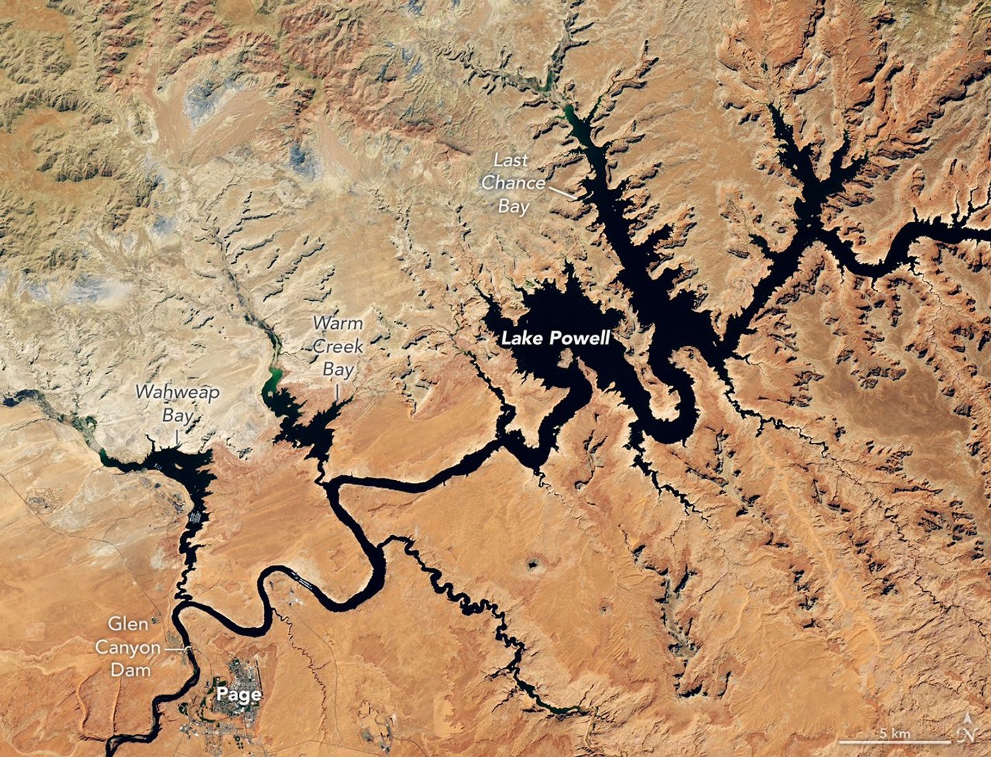 Water in Lake Powell is at record-low levels and looks like a wide river winding through a light orange desert landscape. The Glen Canyon Dam and the town of Page, Arizona, are visible in the lower left.