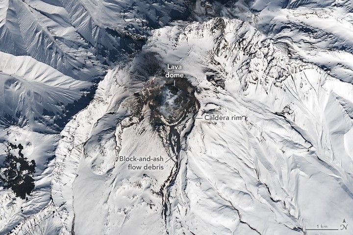 The lava dome appears as a dark patch within the snowy caldera. Dark channels with volcanic deposits are visible draining to the south and west.