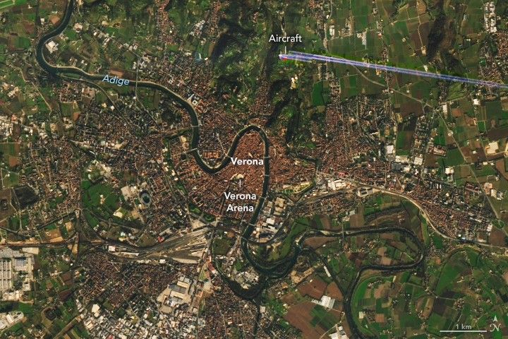 Satellite image of Verona, Italy, showing the historic city center in reddish-brown tones with dense urban development, surrounded by green agricultural fields. A river curves through the upper left, and a labeled marker highlights the ancient Roman amphitheater near the center.