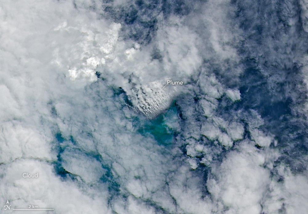 Only small patches of the ocean are visible through puffy, white volcanic plumes streaming from the center of the scene. Clouds not directly related to the eruption are visible throughout much of the image.  A false-color inset box shows the infrared signature of the eruption as a series of red dots near the volcanic plumes.