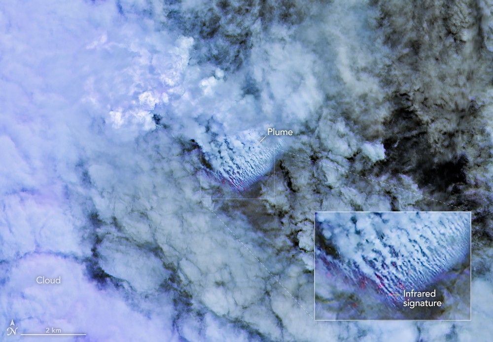 Only small patches of the ocean are visible through puffy, white volcanic plumes streaming from the center of the scene. Clouds not directly related to the eruption are visible throughout much of the image. A false-color inset box shows the infrared signature of the eruption as a series of red dots near the volcanic plumes.