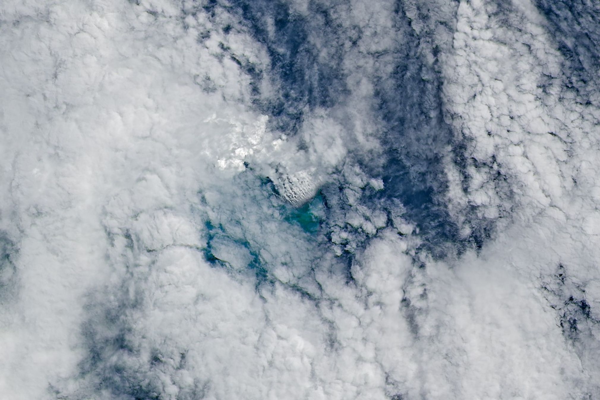 Only small patches of the ocean are visible through puffy, white volcanic plumes streaming from the center of the scene. Clouds not directly related to the eruption are visible throughout much of the image. A false-color inset box shows the infrared signature of the eruption as a series of red dots near the volcanic plumes.