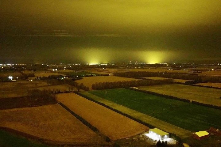 A photo taken at night from above ground level shows rectangular farm fields beneath a sky that is glowing yellow. Two very bright areas on the horizon are emitting beams of light toward the sky. Other, much smaller white dots of light are scattered across the land.