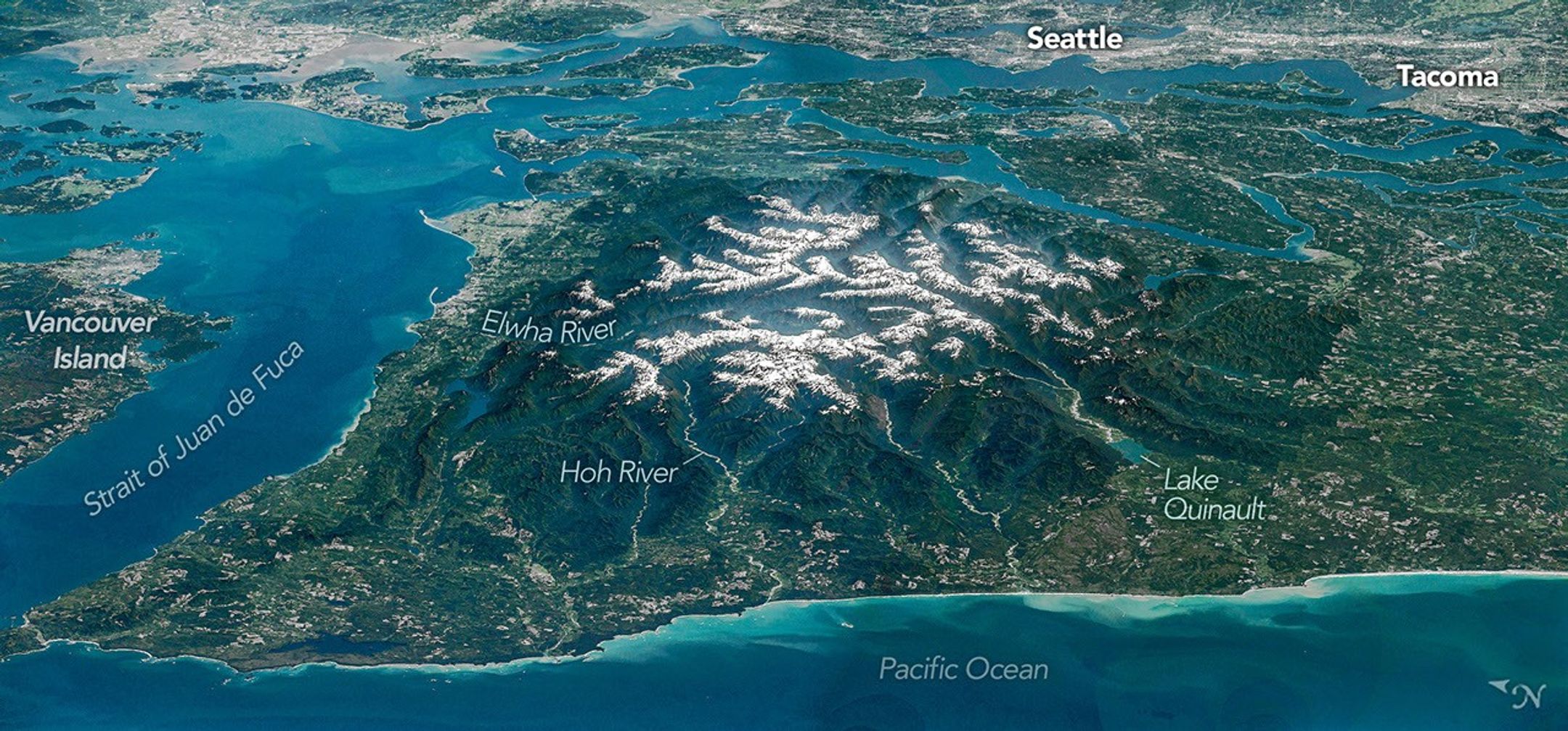The Olympic Peninsula, viewed at an angle from above, features snow-capped mountains surrounded by deep, forested river valleys. Islands in Puget Sound and developed areas including Seattle and Tacoma appear across the top of the photo.