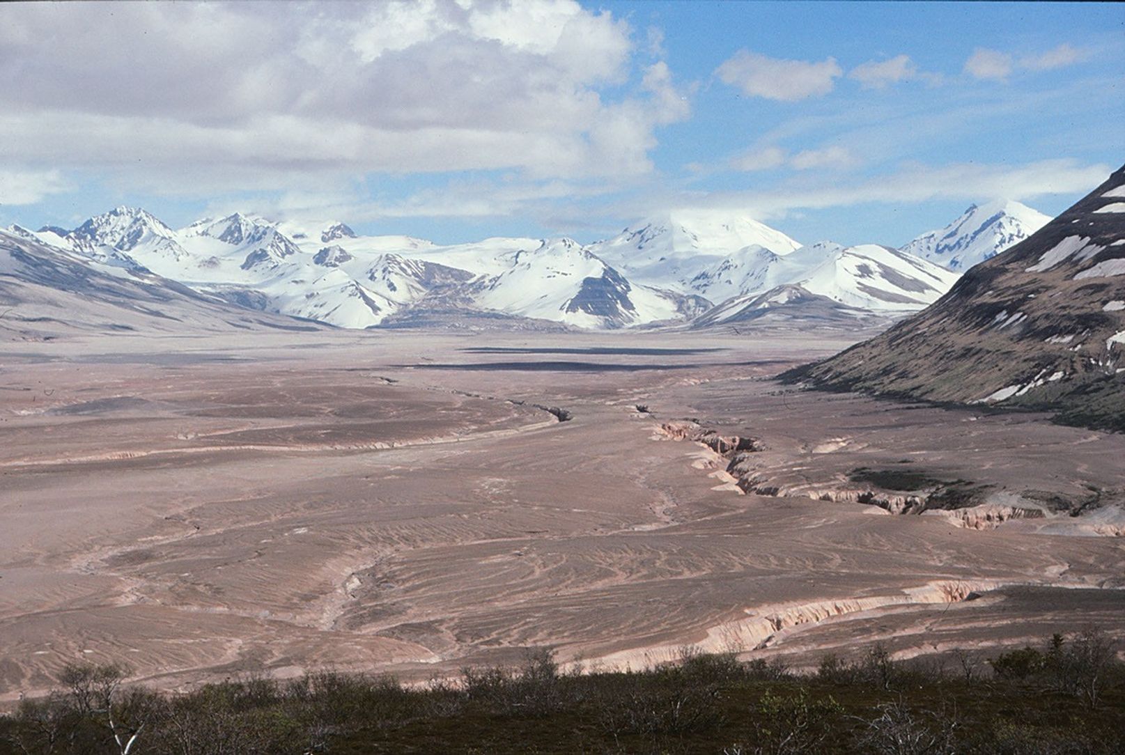 This photo shows a broad valley filled with tan-colored volcanic material in the foreground and snowy mountains in the background. The ashy volcanic deposits create a mostly flat valley floor, except where steep-sided chasms formed by erosion run through it.
