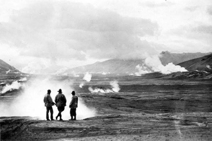 Three people stand in the foreground of this black-and-white photo, silhouetted by steam coming from a fumarole. A dark, barren landscape dotted with plumes of rising steam extends into the background.