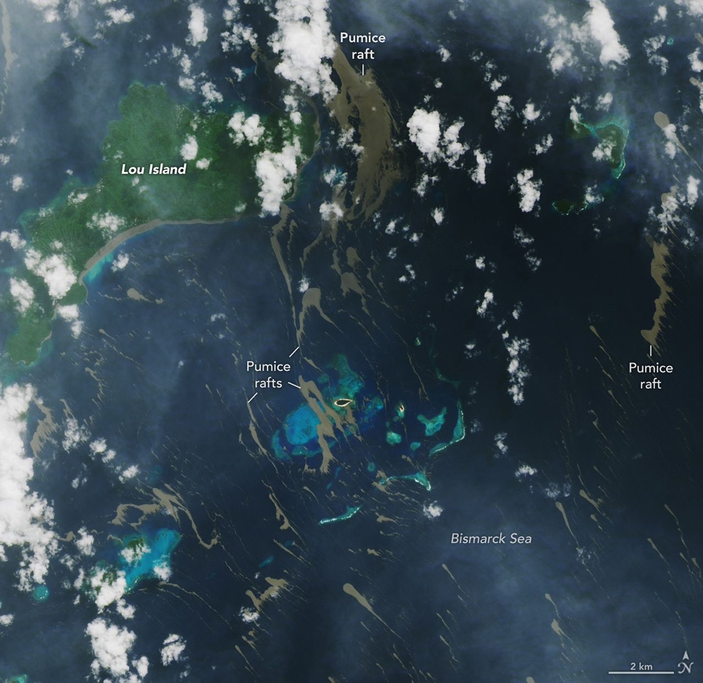 Bands of tan-colored pumice float on the Bismarck Sea near several islands and bright-blue shallow areas. The material lines the southern coast of the largest island.