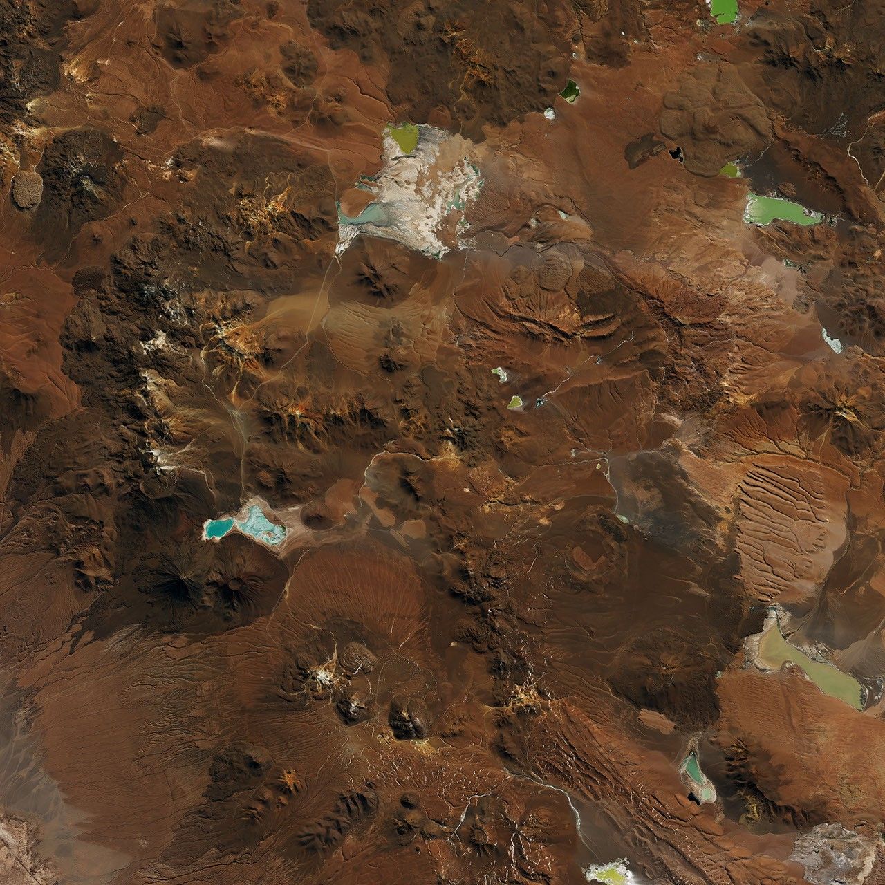 A plateau in the Chilean Andes is surrounded by stratovolcanoes and lava domes. The dry, mostly brown landscape has a lighter brown patch near the center, marking the radio telescope array.