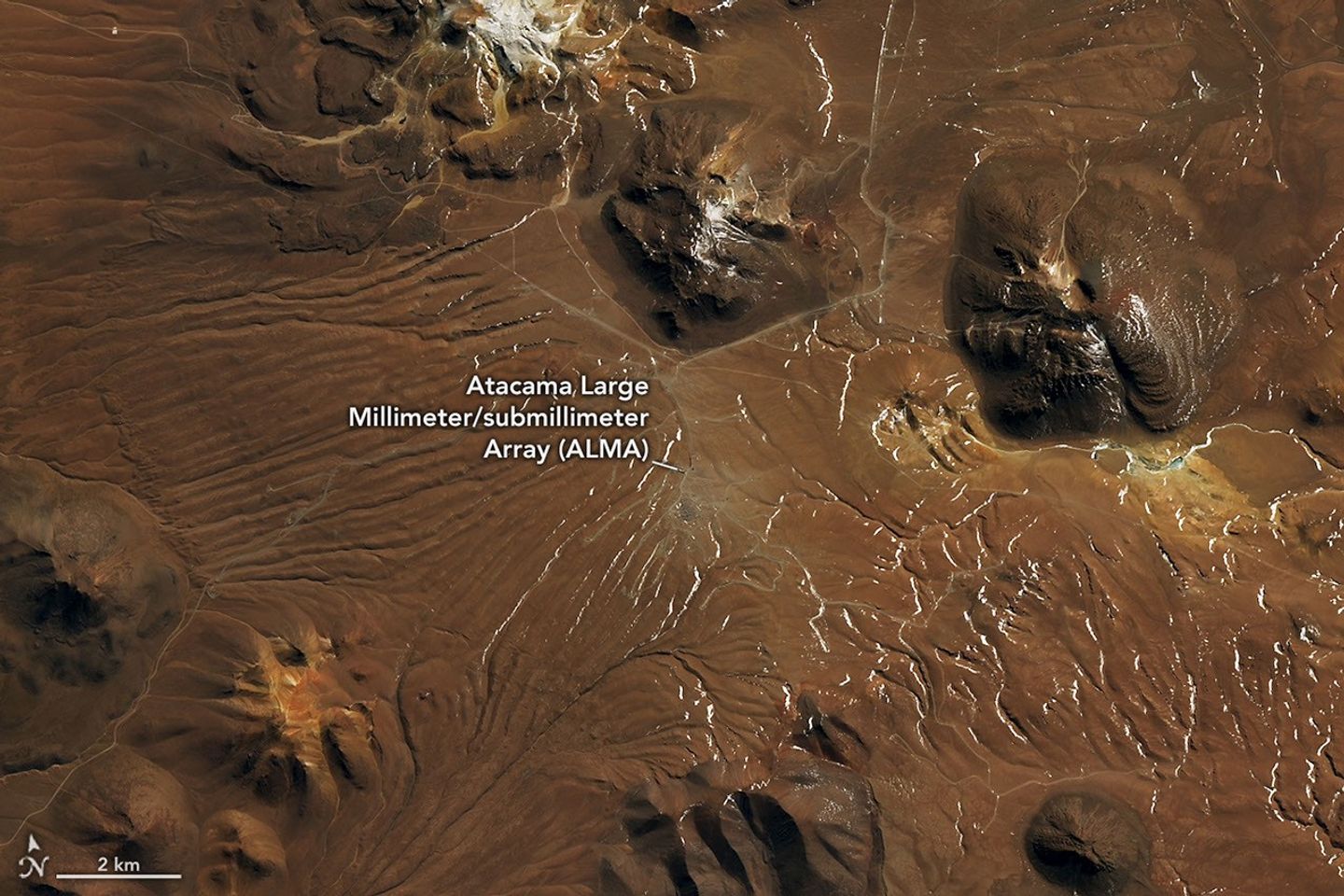 A plateau in the Chilean Andes is surrounded by stratovolcanoes and lava domes. The dry, mostly brown landscape has a lighter brown patch near the center, marking the radio telescope array.