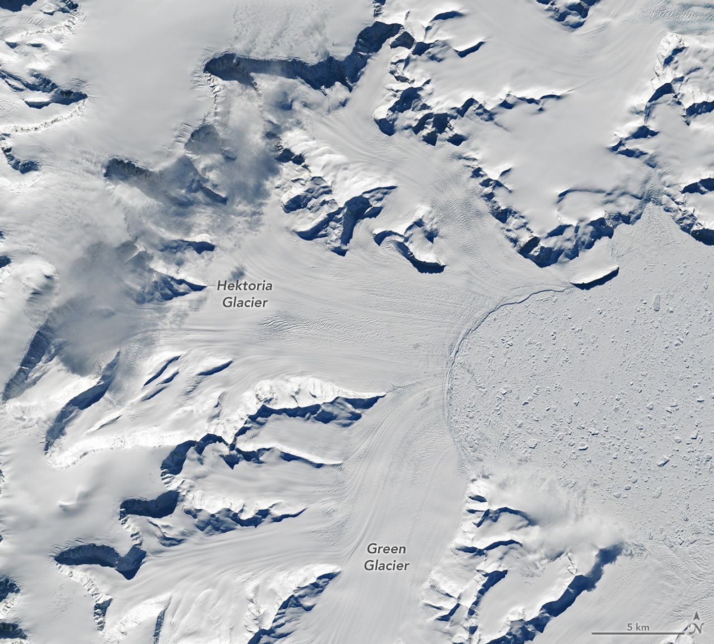 Glaciers flow from cliffs on the left and bottom sides of the image to a frozen bay on the right where chunks of sea ice are visible.