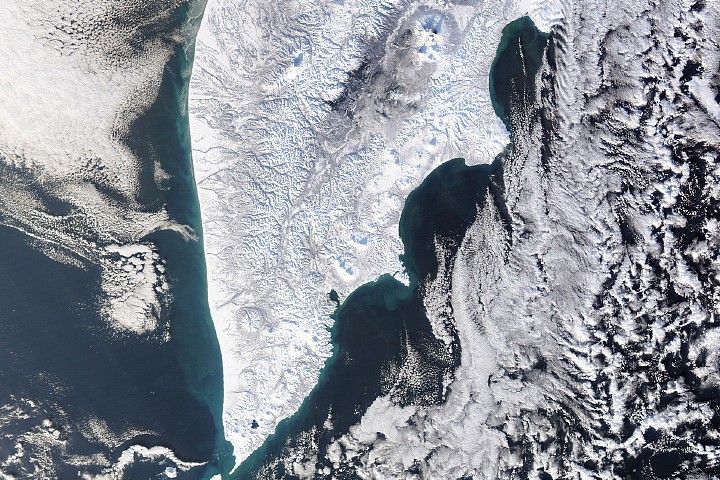 A thick layer of white snow blankets the Kamchatka Peninsula. Layers of clouds surround the peninsula, framing it but leaving its coastlines and a narrow portion of ocean visible around it. On land, several large, circular volcanoes dot the rugged landscape.