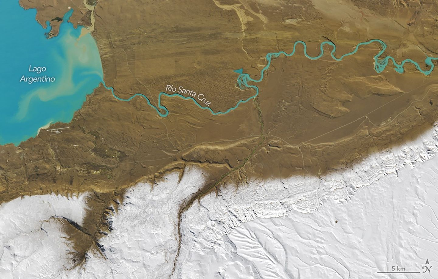 The outlet of bright blue Lago Argentino meanders across an otherwise dry, brown valley from left to right. The bottom third of the image is white, where snow covers higher elevations.