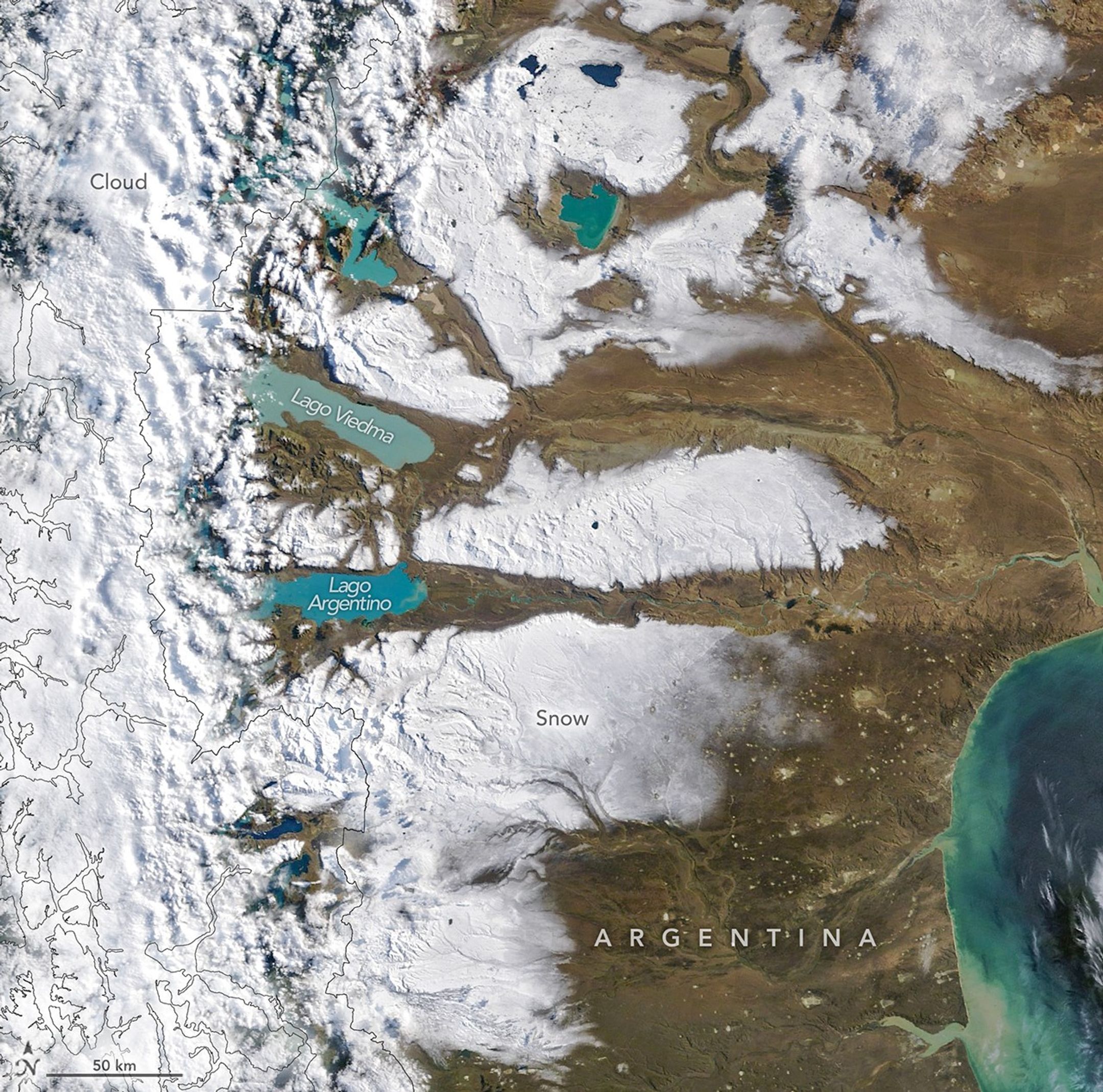 Large areas of snow cover the arid plains of southern Argentina. Several large lakes containing water in shades of milky blue are visible, while the Andes are veiled by clouds along the left side.