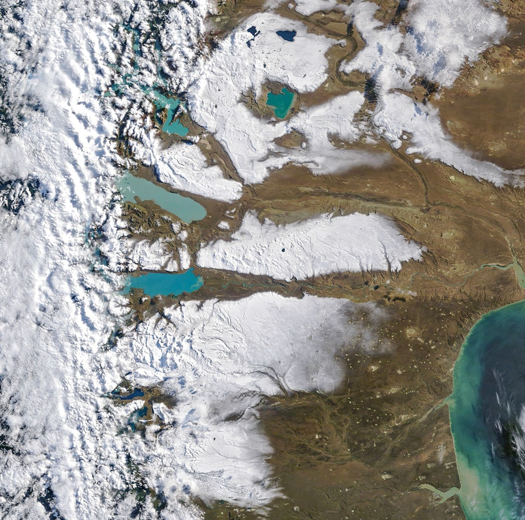 Large areas of snow cover the arid plains of southern Argentina. Several large lakes containing water in shades of milky blue are visible, while the Andes are veiled by clouds along the left side.