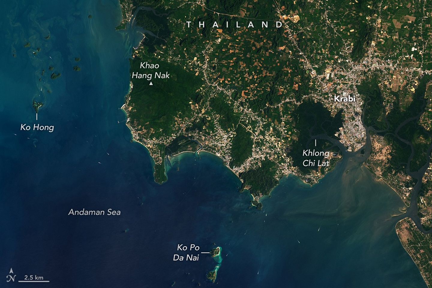 Alt text: A wide view of Krabi Province shows offshore islands and boats in blue water and inland areas with a mix of gray urban development, brown farmland, and green vegetation.
