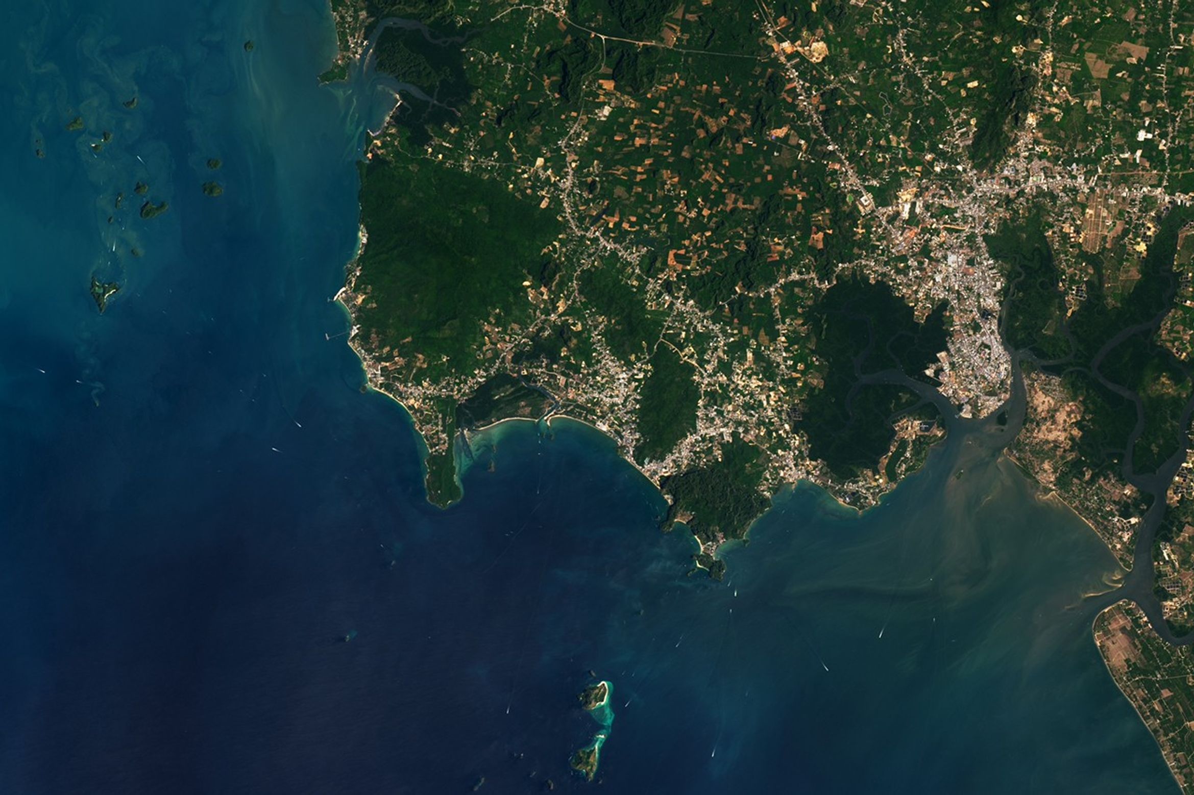 Alt text: A wide view of Krabi Province shows offshore islands and boats in blue water and inland areas with a mix of gray urban development, brown farmland, and green vegetation.