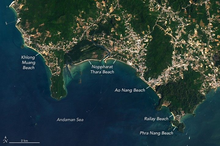 Strips of sandy beach line the coast of Krabi Province, Thailand, separating blue ocean water from inland greenery and urban areas.