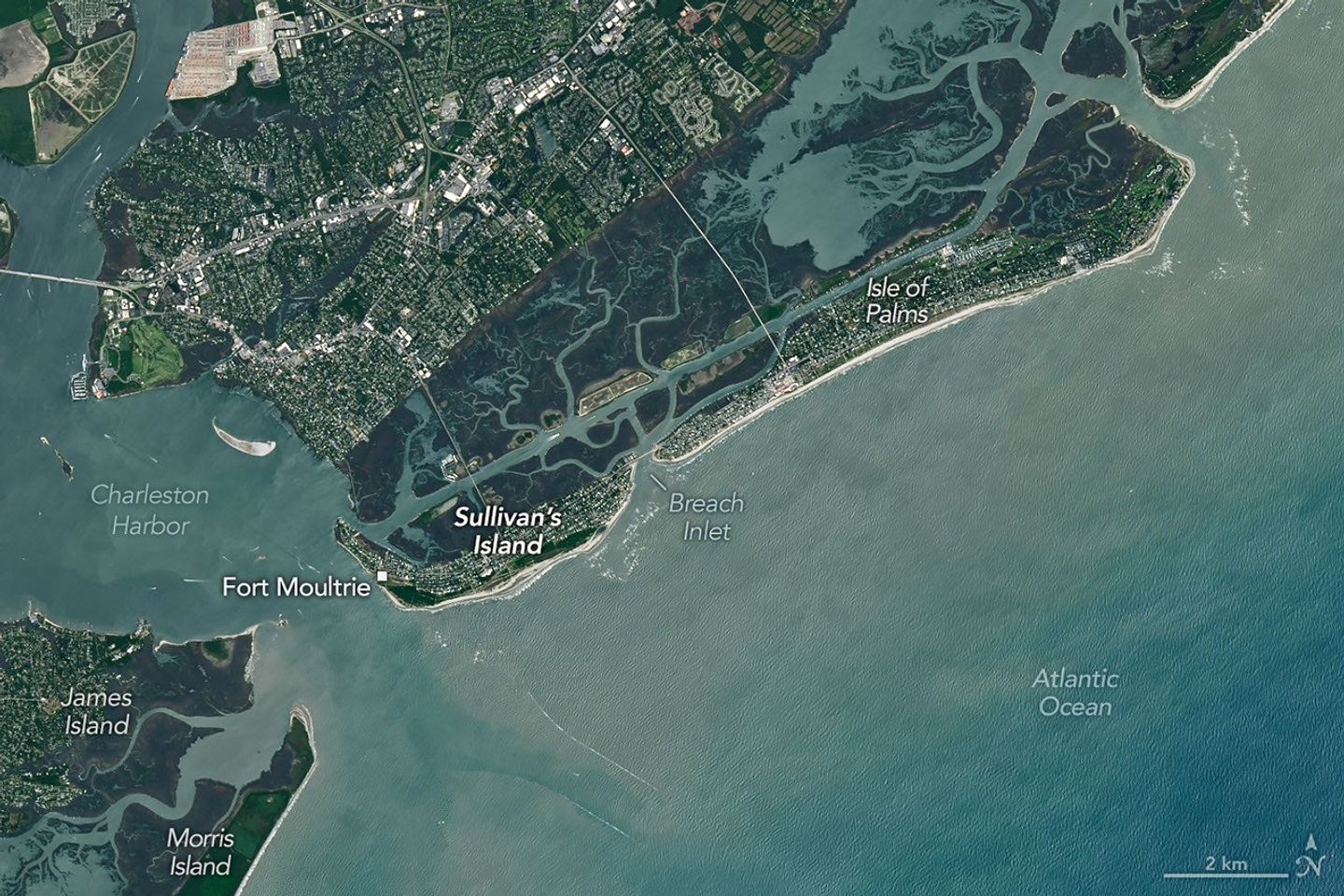 A satellite image shows Sullivan’s Island, South Carolina, with a sandy shoreline along the Atlantic Ocean to the south, development across the interior, and green marsh and tidal inlets along its north side.