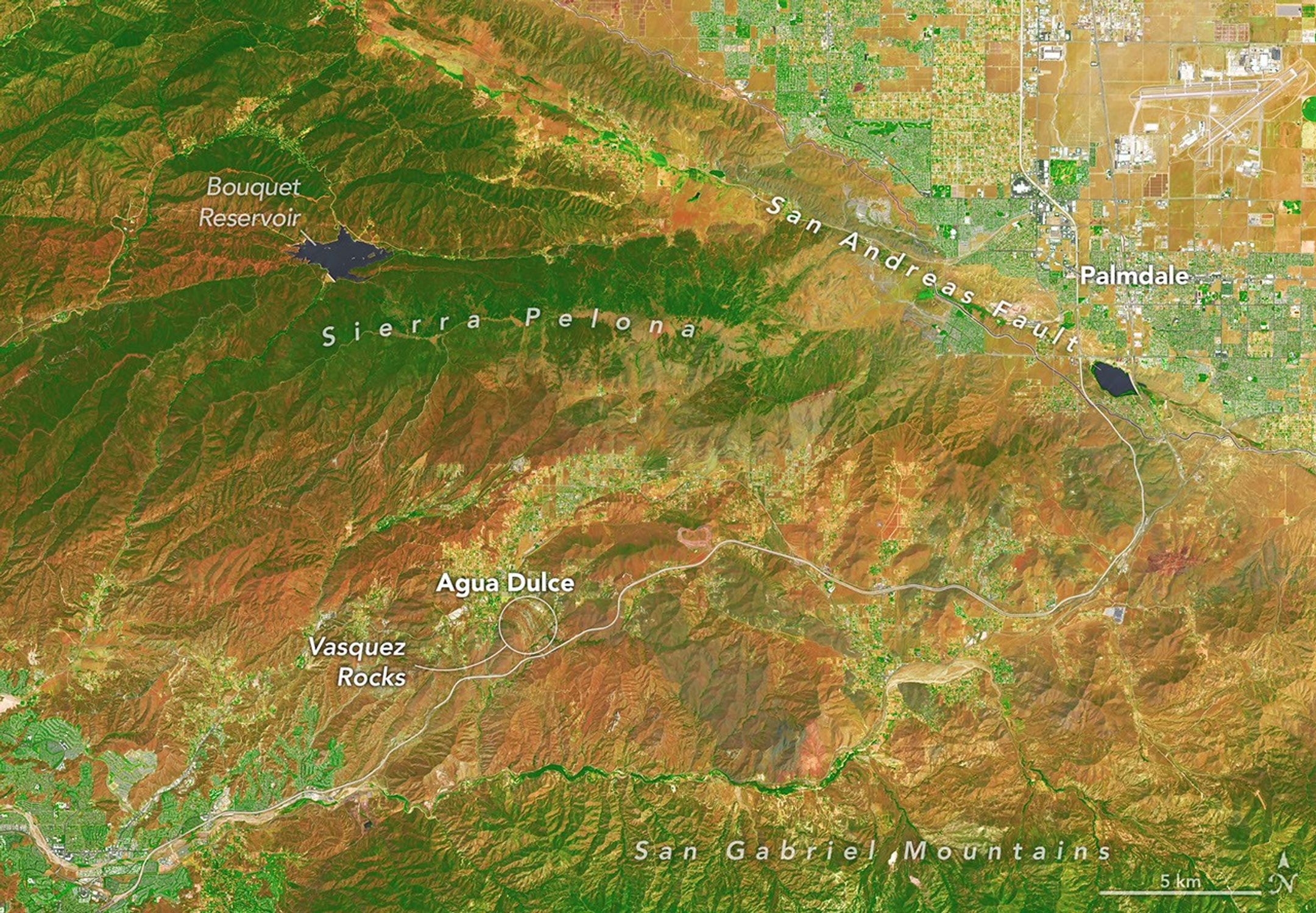 In this false-color image, areas with more extensive vegetation on the Sierra Pelona retain more moisture than the hills surrounding Agua Dulce, making the Sierra Pelona appear dark green in comparison to the rusty brown coloration of the lower hills. Vasquez Rocks appears as a patch of curved gray stripes near the center of the image.