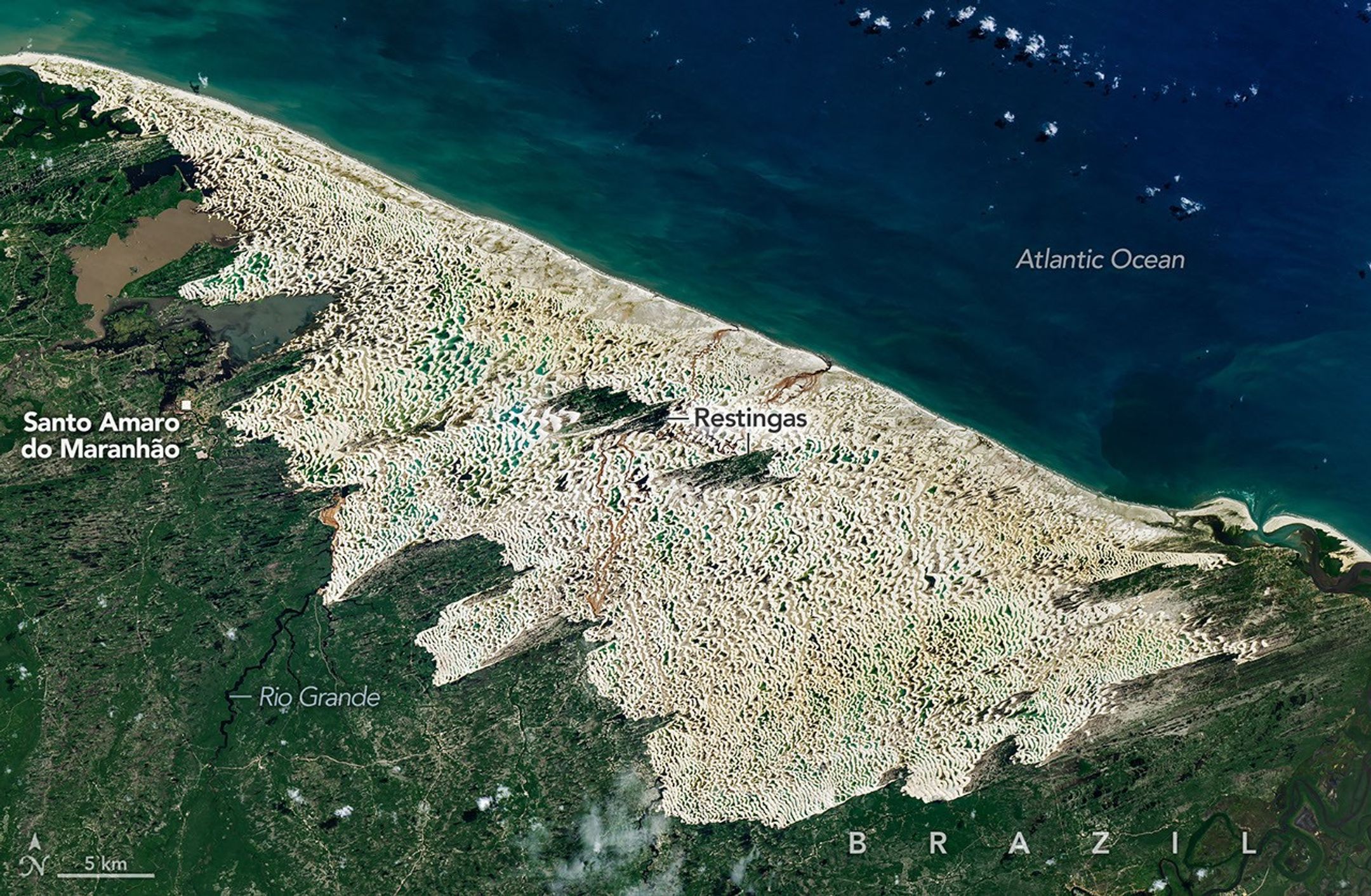Bright white sand dunes stretch for dozens of kilometers between the dark blue Atlantic Ocean and green coastal vegetation. Blue and green freshwater lagoons fill low areas between the dunes, while dark-green vegetated patches lie within the dune field.