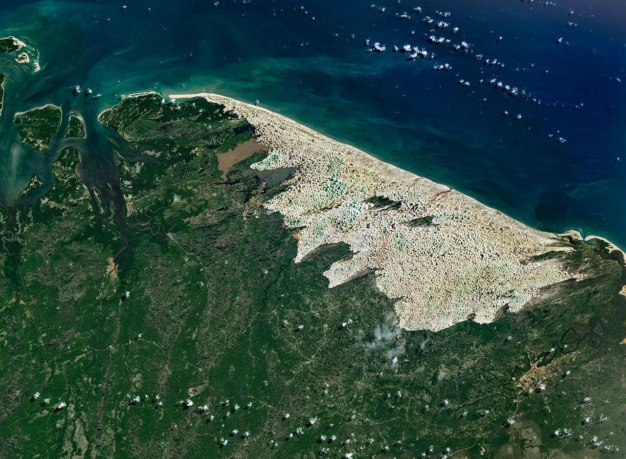 Bright white sand dunes stretch for dozens of kilometers between the dark blue Atlantic Ocean and green coastal vegetation. Blue and green freshwater lagoons fill low areas between the dunes, while dark-green vegetated patches lie within the dune field.