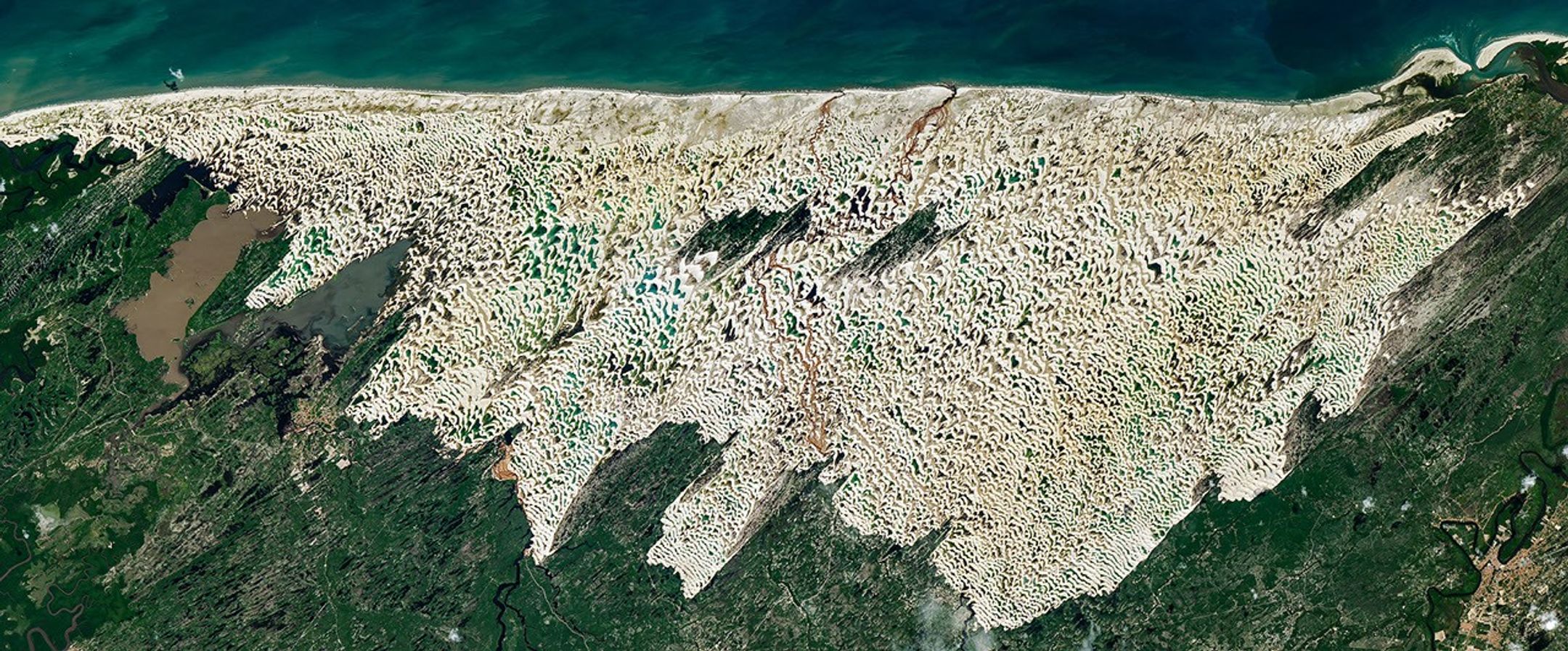Bright white sand dunes stretch for dozens of kilometers between the dark blue Atlantic Ocean and green coastal vegetation. Blue and green freshwater lagoons fill low areas between the dunes, while dark-green vegetated patches lie within the dune field.
