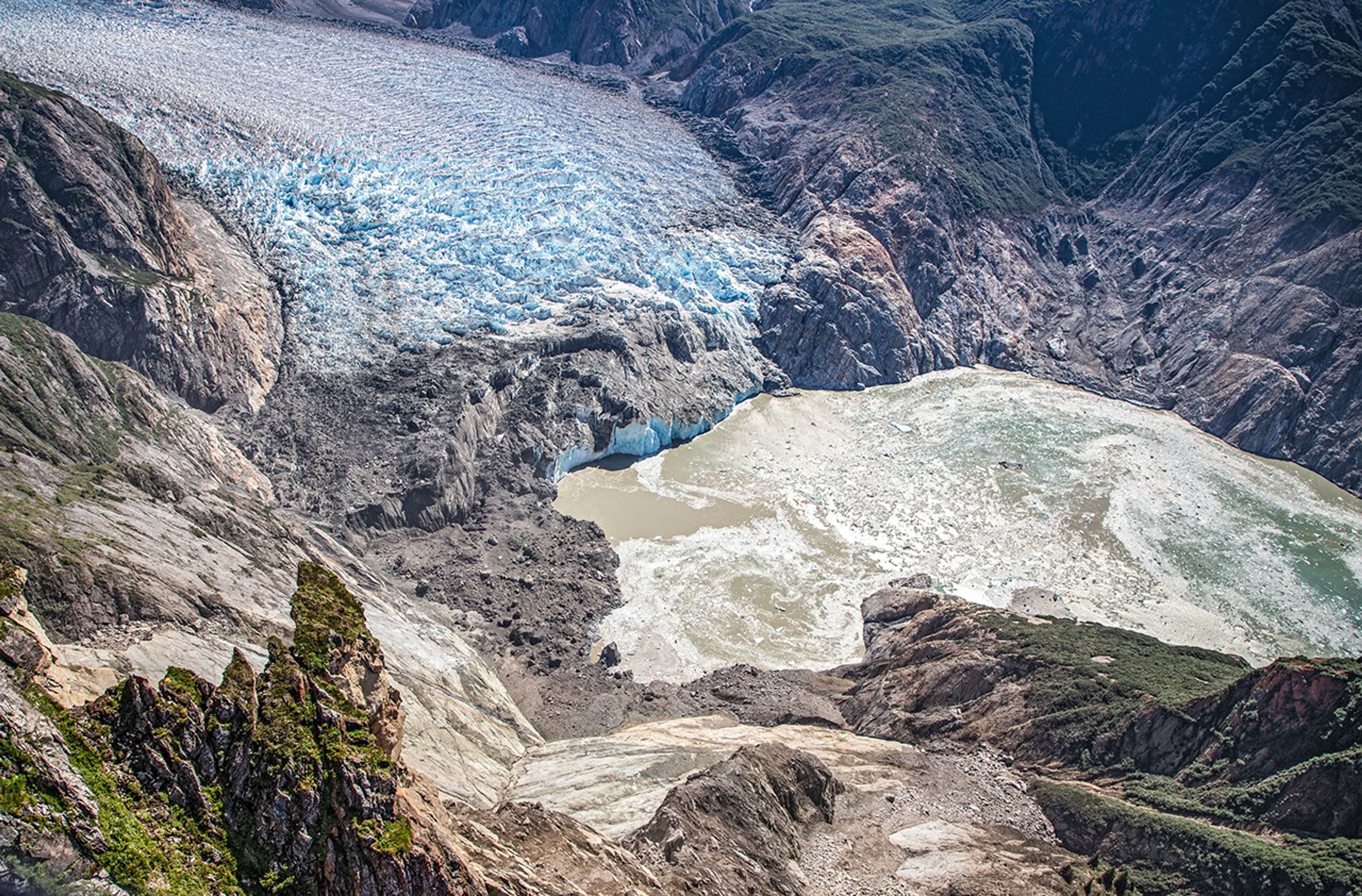 An aerial view shows a glacier ending in a fjord with small icebergs in the water. A recent landslide scars the foreground fjord wall, while a tsunami has stripped vegetation from the opposite wall.